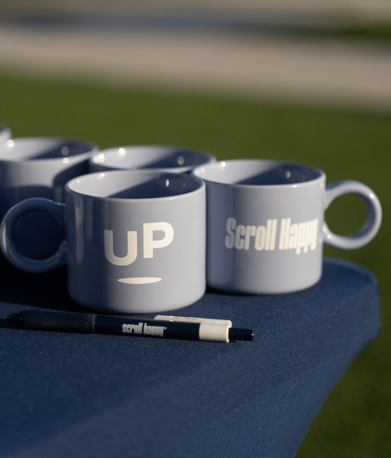 Periwinkle UP mugs and a Scroll Happy pen on a navy table outdoors