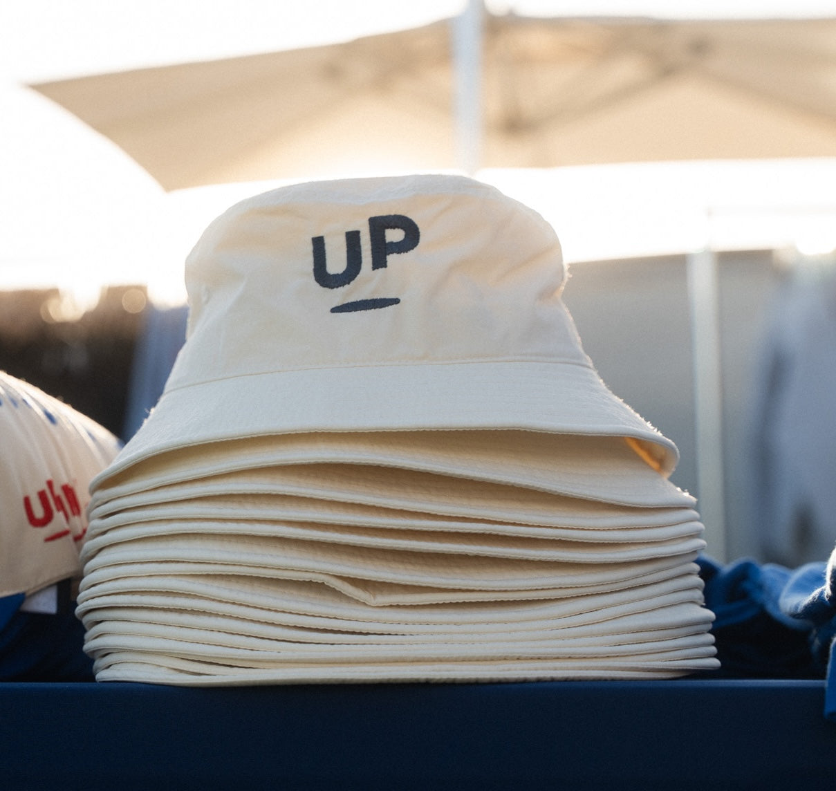 Cream UP bucket hat with the navy embroidered UP logo, stacked on a table