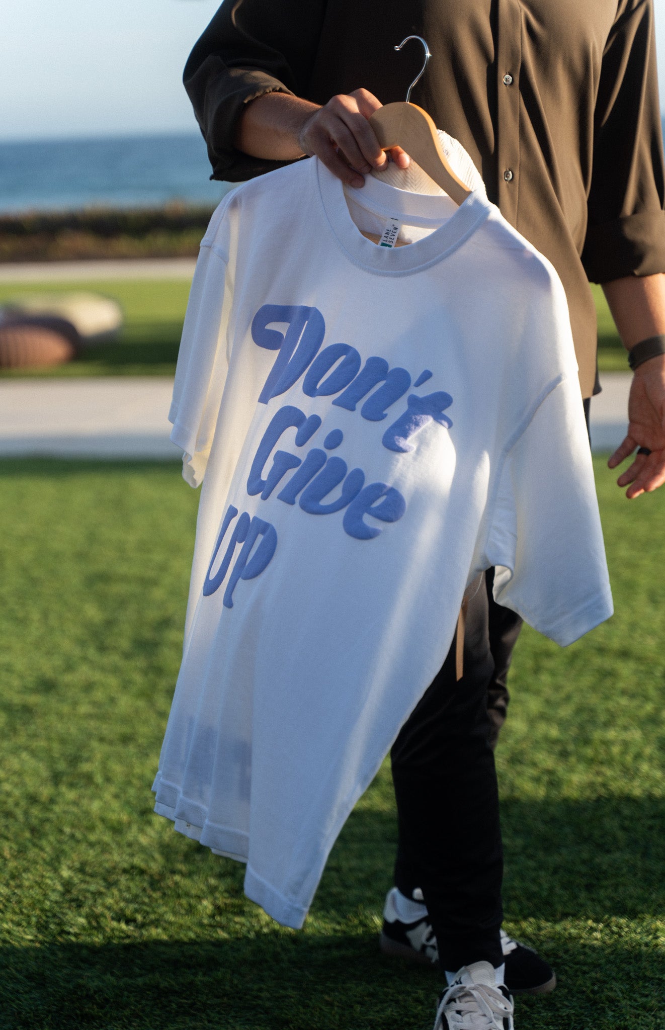 Don't Give UP tee held on a hanger outdoors, blue script lettering across the front