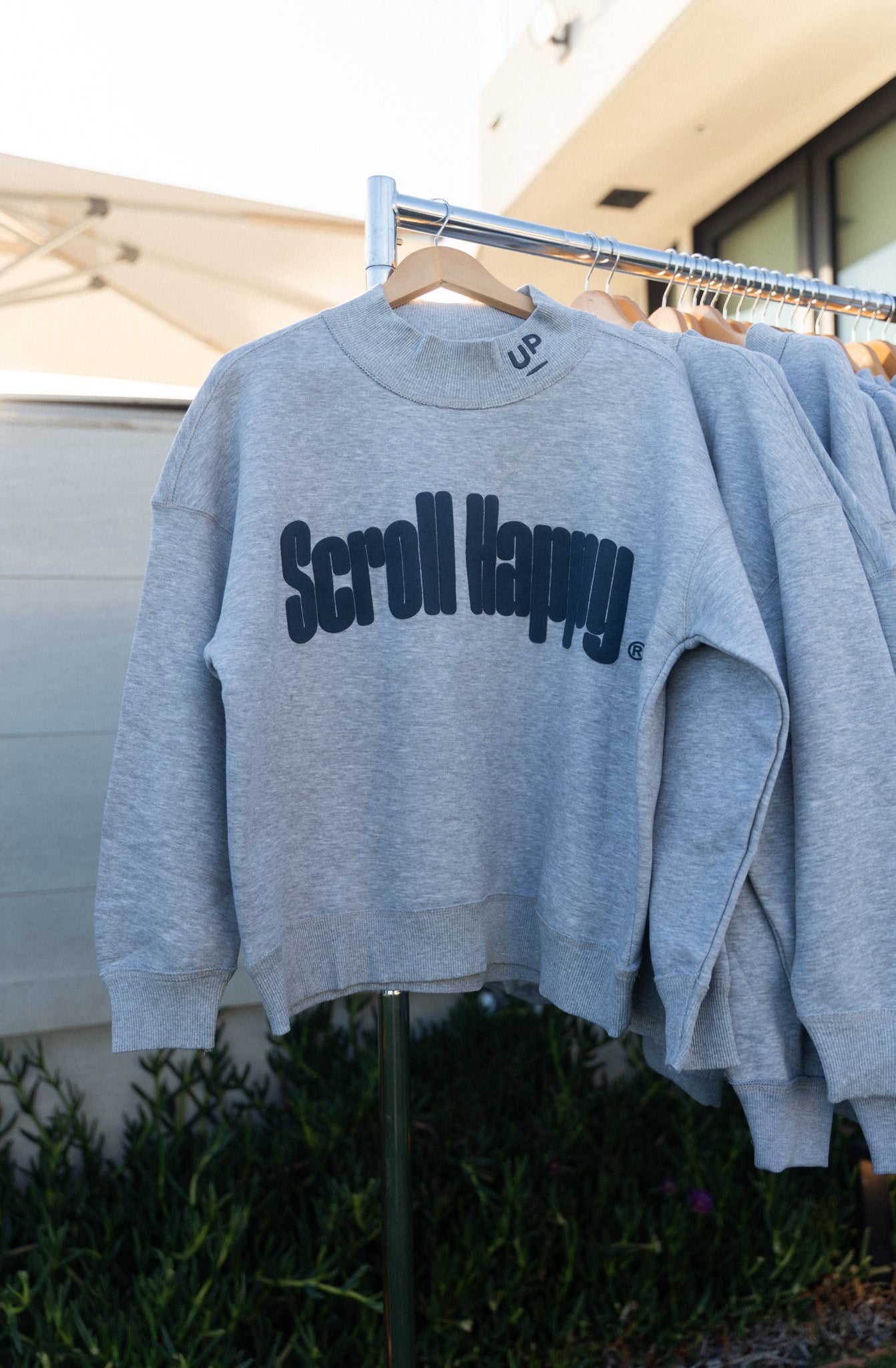 Grey Scroll Happy mock neck sweatshirt hanging on a rack outdoors